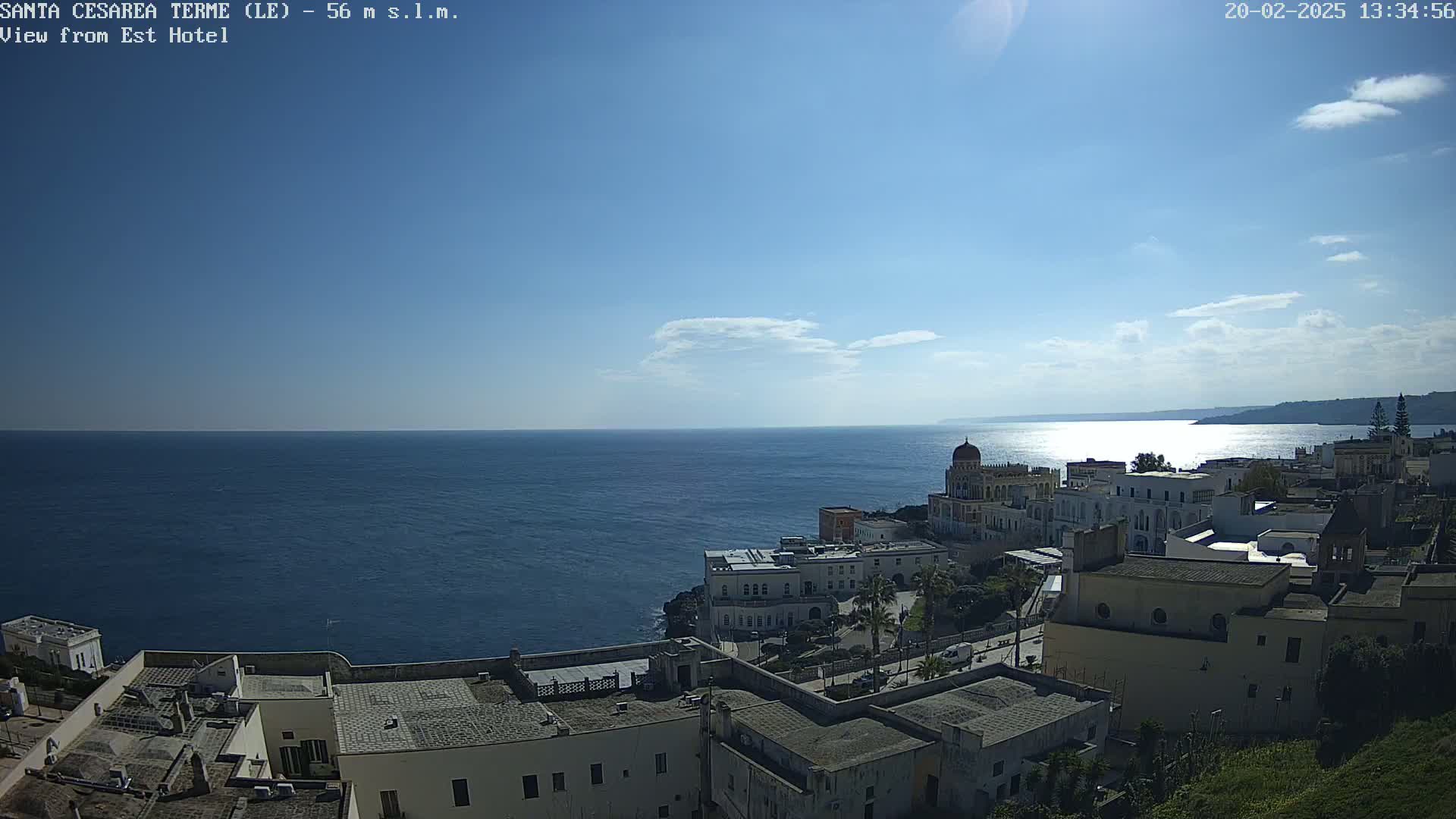 A coastal town with white buildings sits on a cliff overlooking a calm, blue sea under a mostly sunny sky.