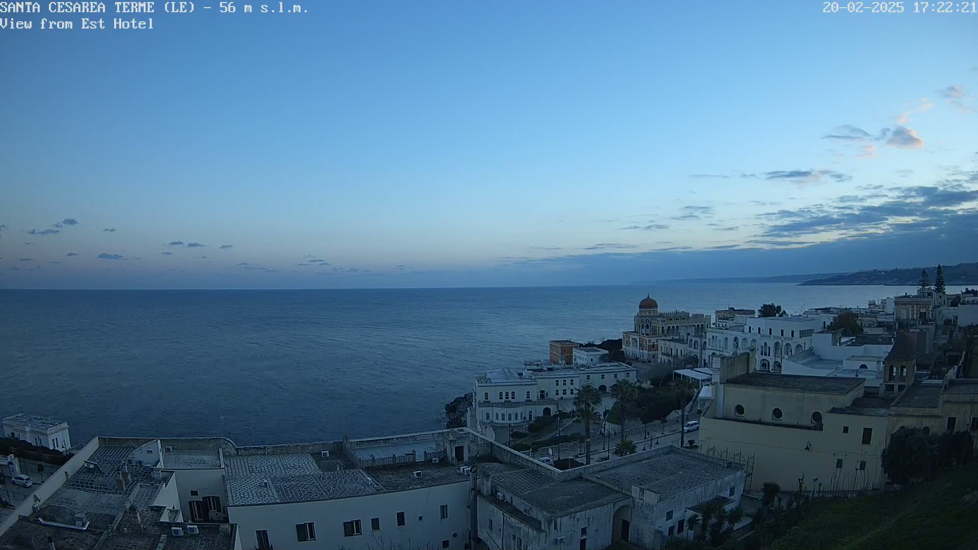 A coastal town with white buildings and a domed structure overlooks a calm, dark-blue sea under a twilight sky with a few clouds.