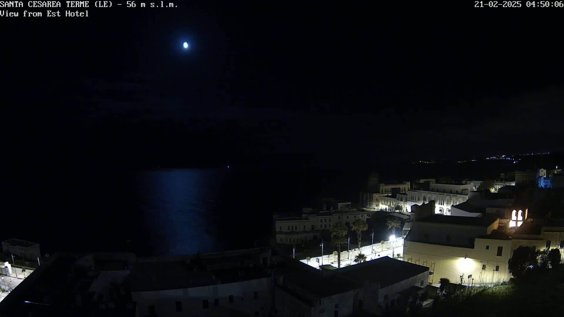 A nighttime, moonlit view of a coastal town with buildings illuminated along a dark body of water.