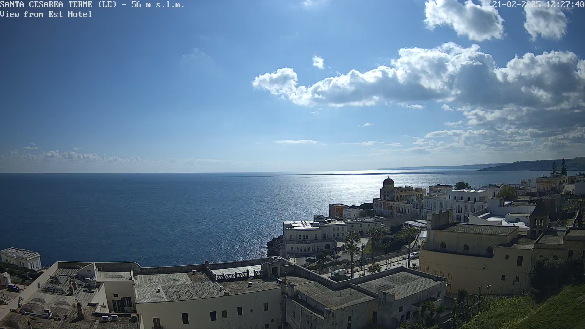 A partly sunny day reveals a coastal town with white buildings overlooking a calm blue sea under a mostly clear sky dotted with fluffy clouds.