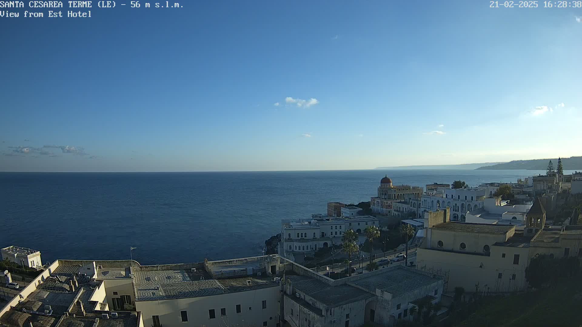 A coastal town with white buildings and a blue-domed structure overlooks a calm, dark-blue sea under a clear, mostly sunny sky.