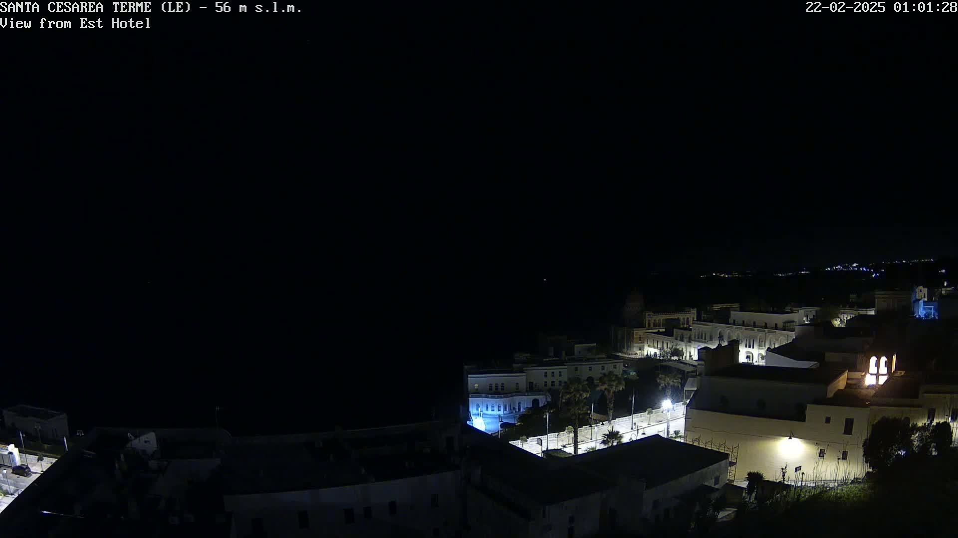A nighttime, high-angle view shows a coastal town with buildings illuminated against a dark sea and sky.