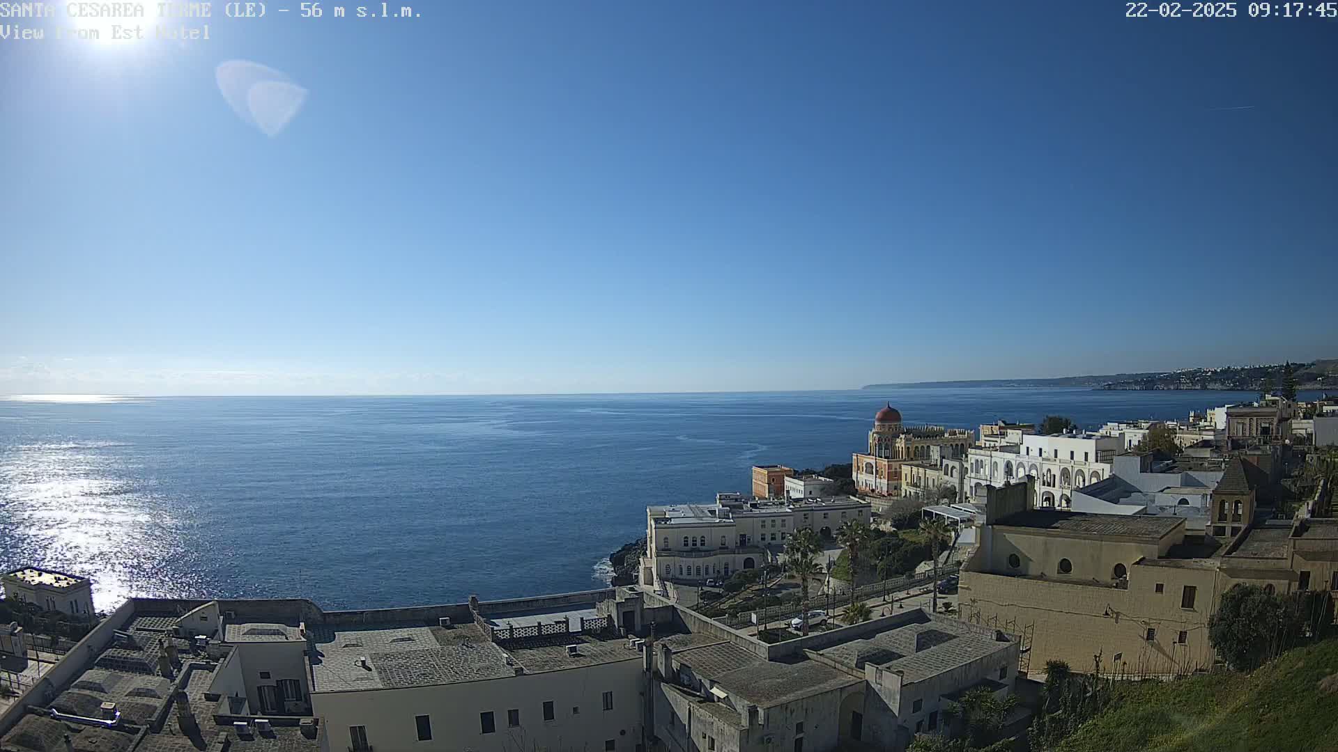 A coastal town with white buildings overlooks a calm, blue sea under a clear, sunny sky.
