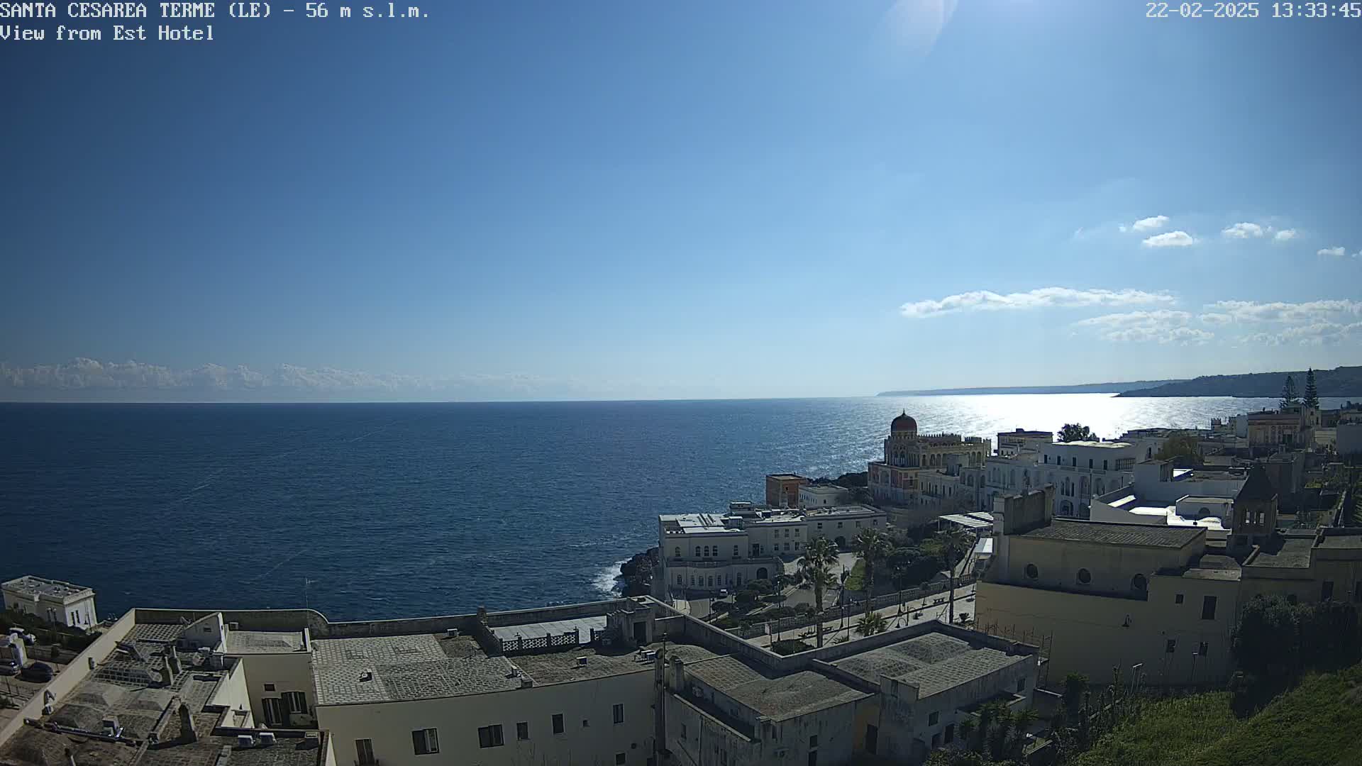 A coastal town with white buildings and a calm blue sea under a mostly clear, sunny sky.