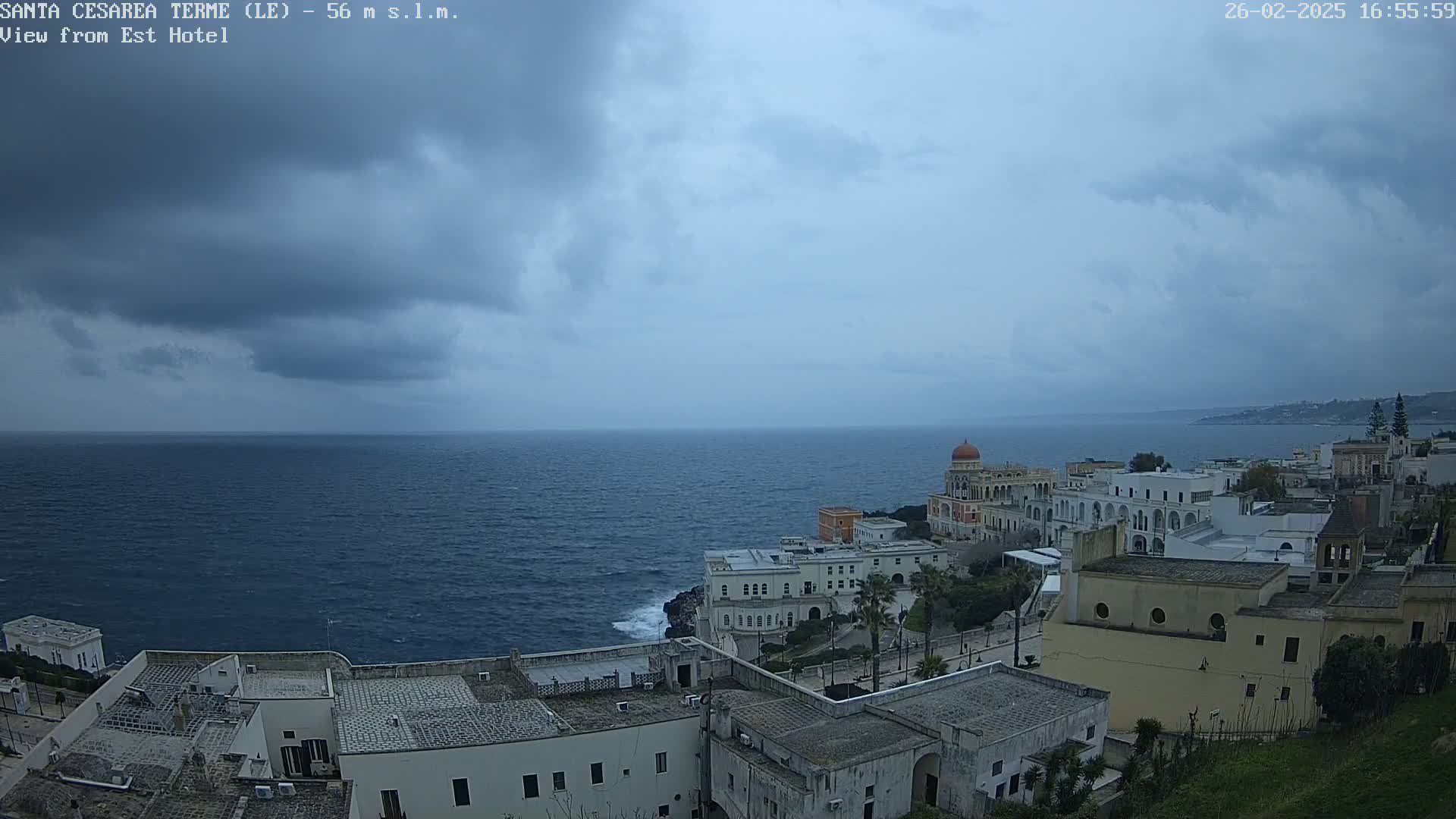 A coastal town with white buildings overlooks a dark blue sea under a cloudy sky.