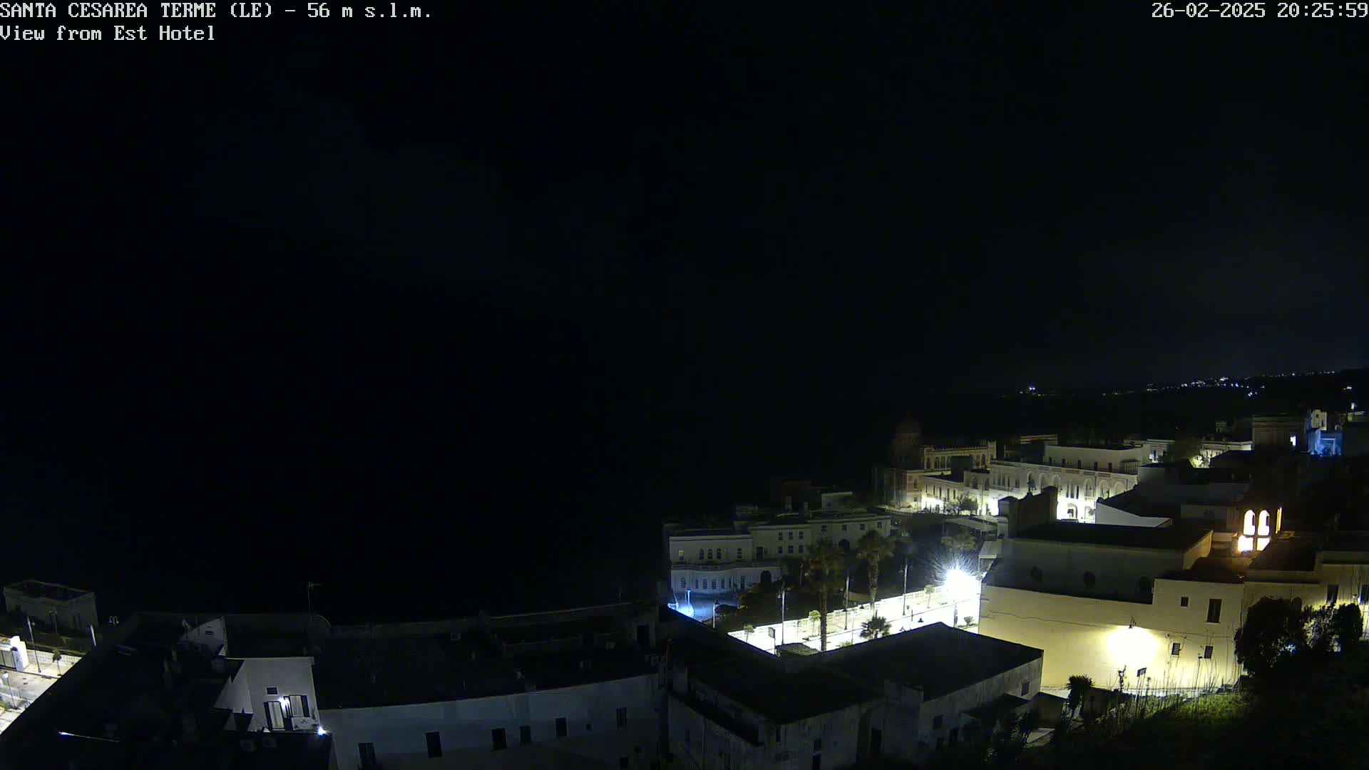 A nighttime, high-angle view of a coastal town with buildings illuminated, under a dark, clear sky.