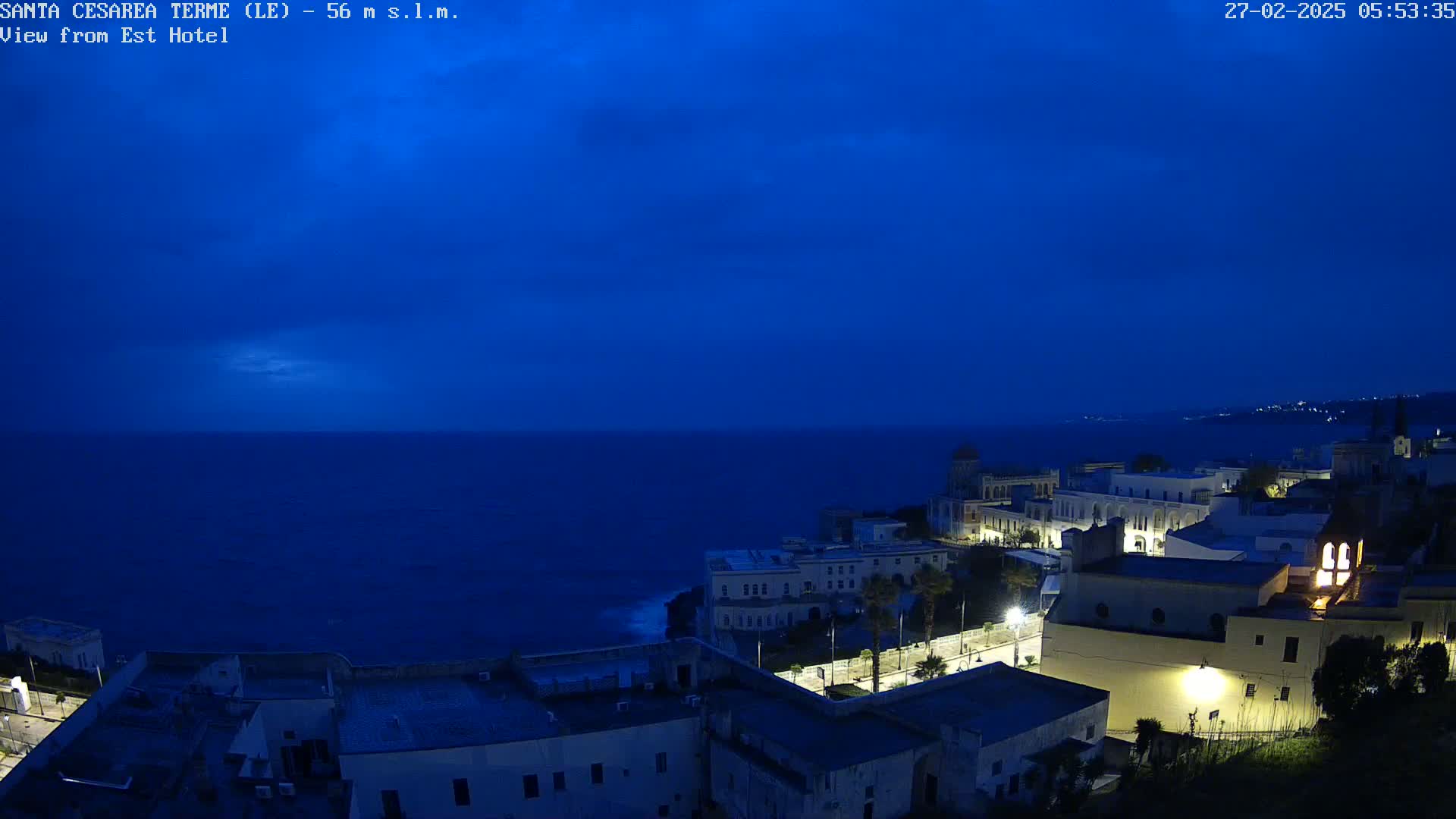 A dark-blue, pre-dawn sky overlooks a coastal town illuminated by lights at night, with a dark blue sea in the background.