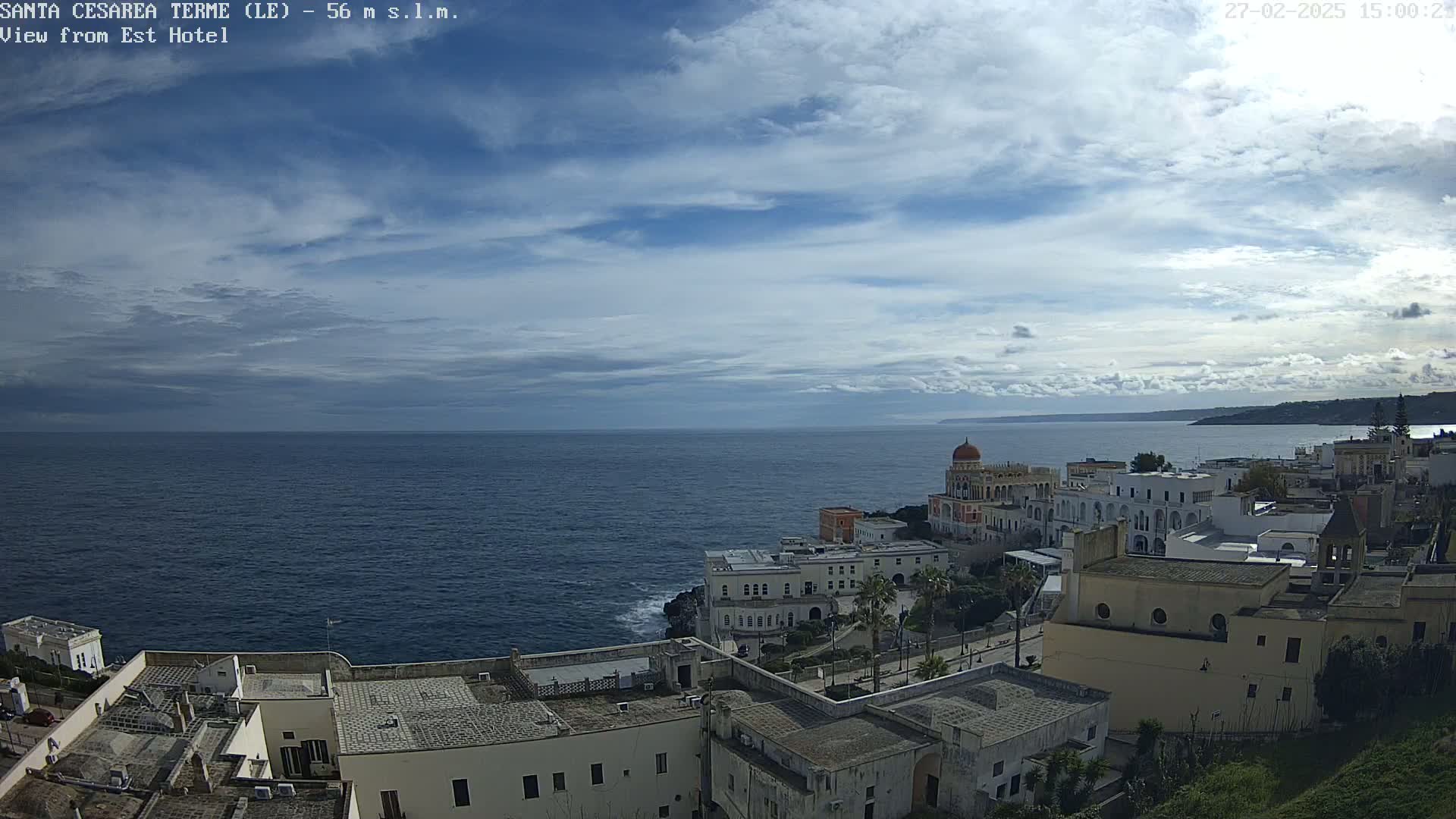 A coastal town with white buildings overlooks a dark blue sea under a partly cloudy sky.
