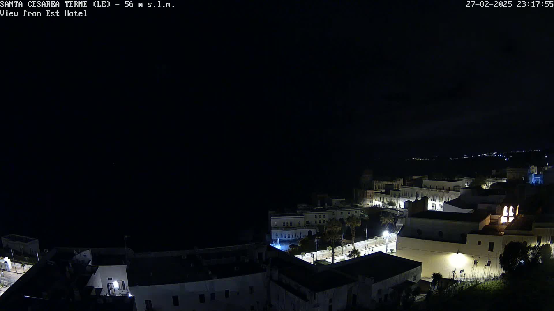 A nighttime, long shot of a coastal town with sparsely lit buildings and a dark sea under a clear, black sky.