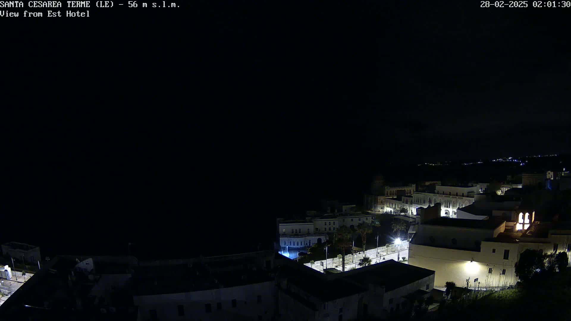 A nighttime, dark, coastal town with sparsely lit buildings is seen from an elevated vantage point under a clear, dark sky.
