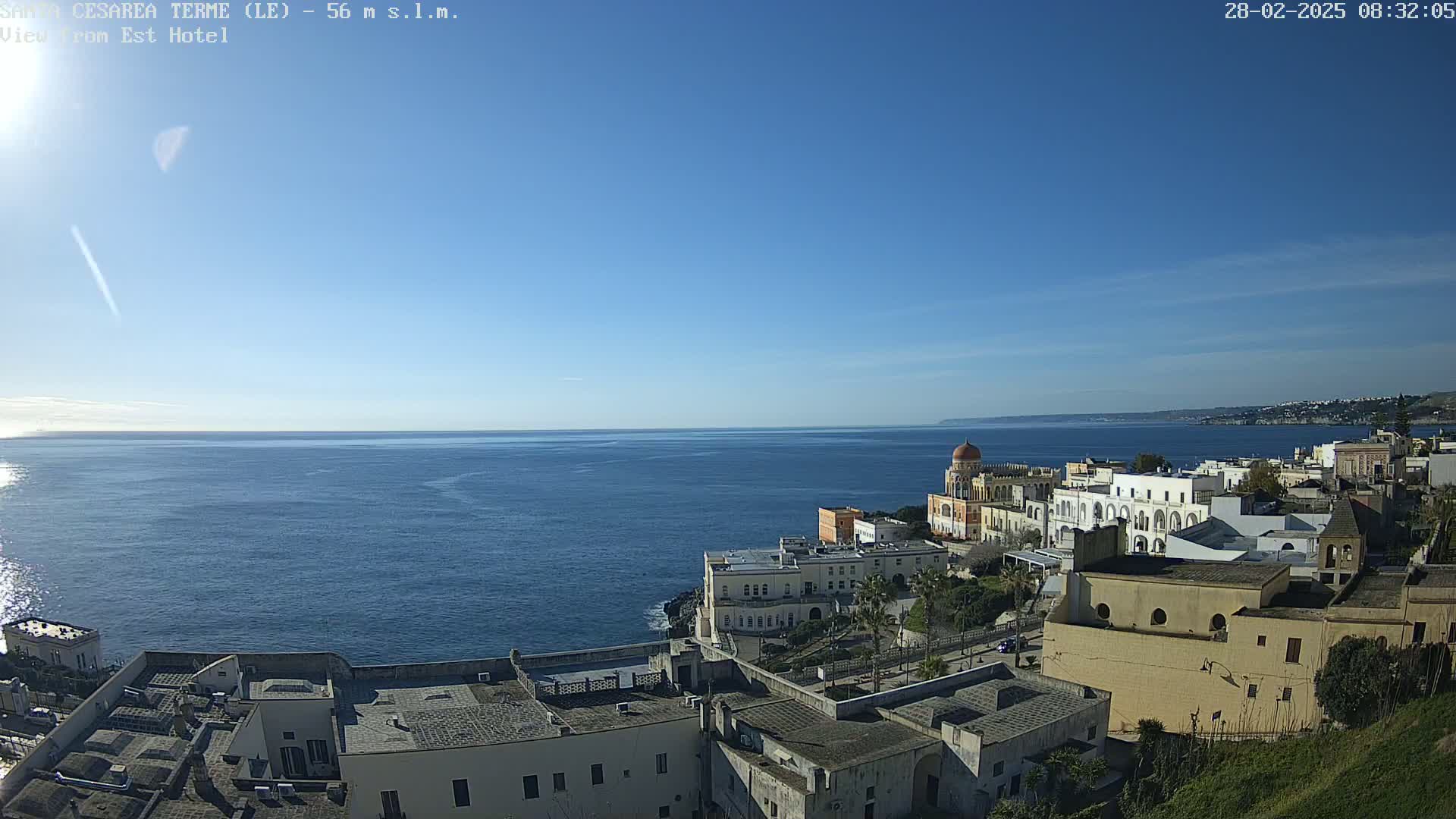 A coastal town with light-colored buildings overlooks a calm, blue sea under a clear, sunny sky.