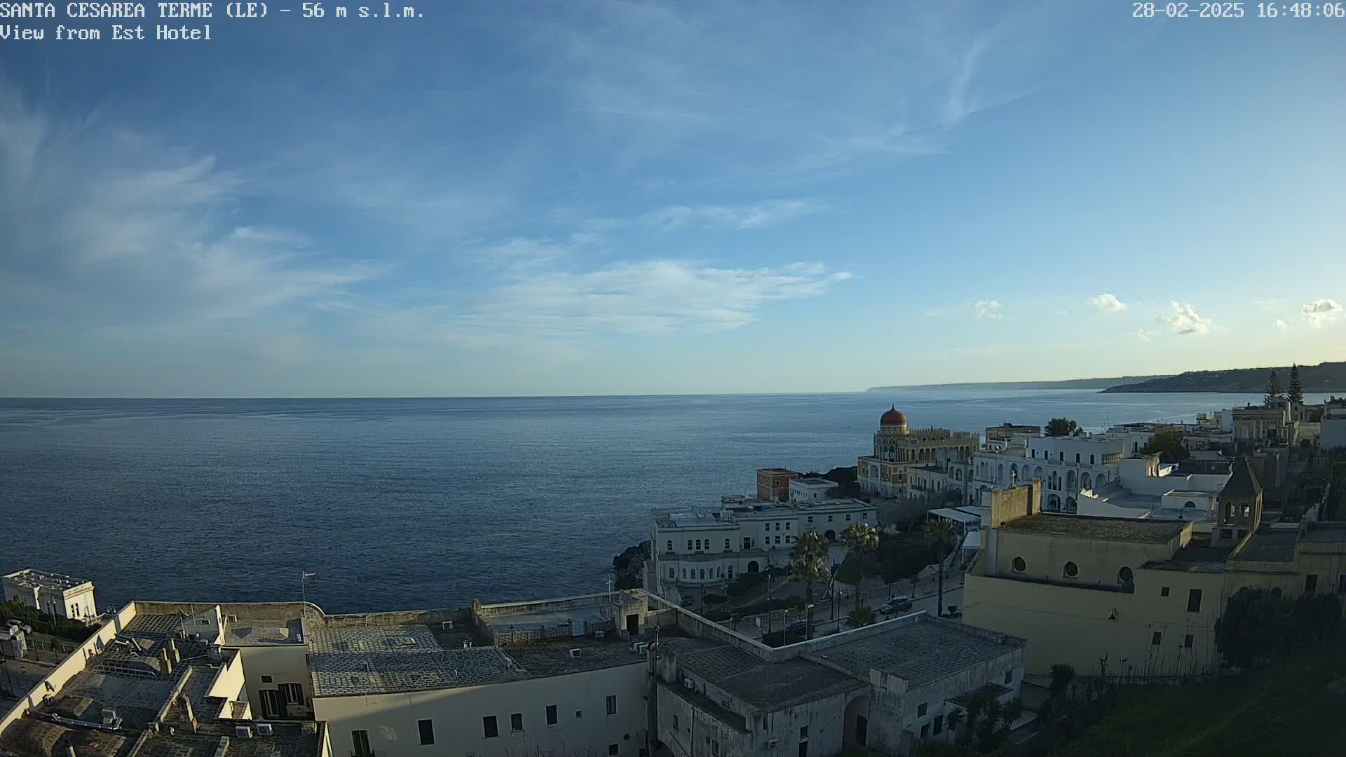 A coastal town with white buildings, a domed structure, and a calm sea under a mostly clear, light blue sky.