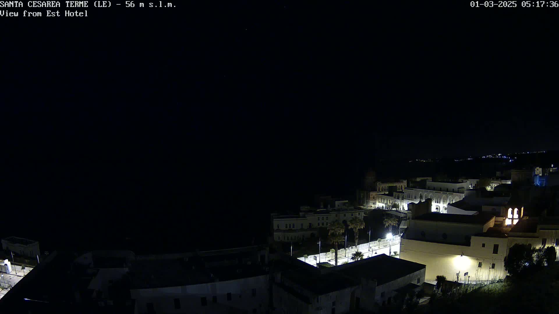 A nighttime, long shot overlooking a coastal town with dimly lit buildings and streets.