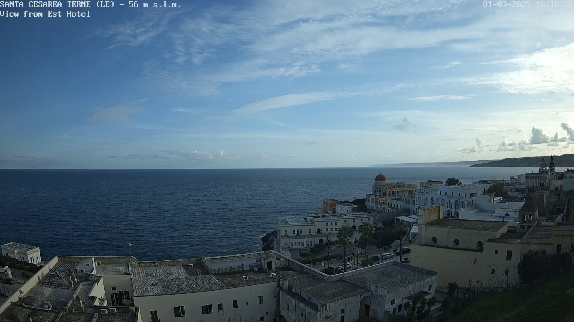 A coastal town with white buildings, a prominent domed structure, and a dark blue sea under a mostly clear sky with a few scattered clouds.