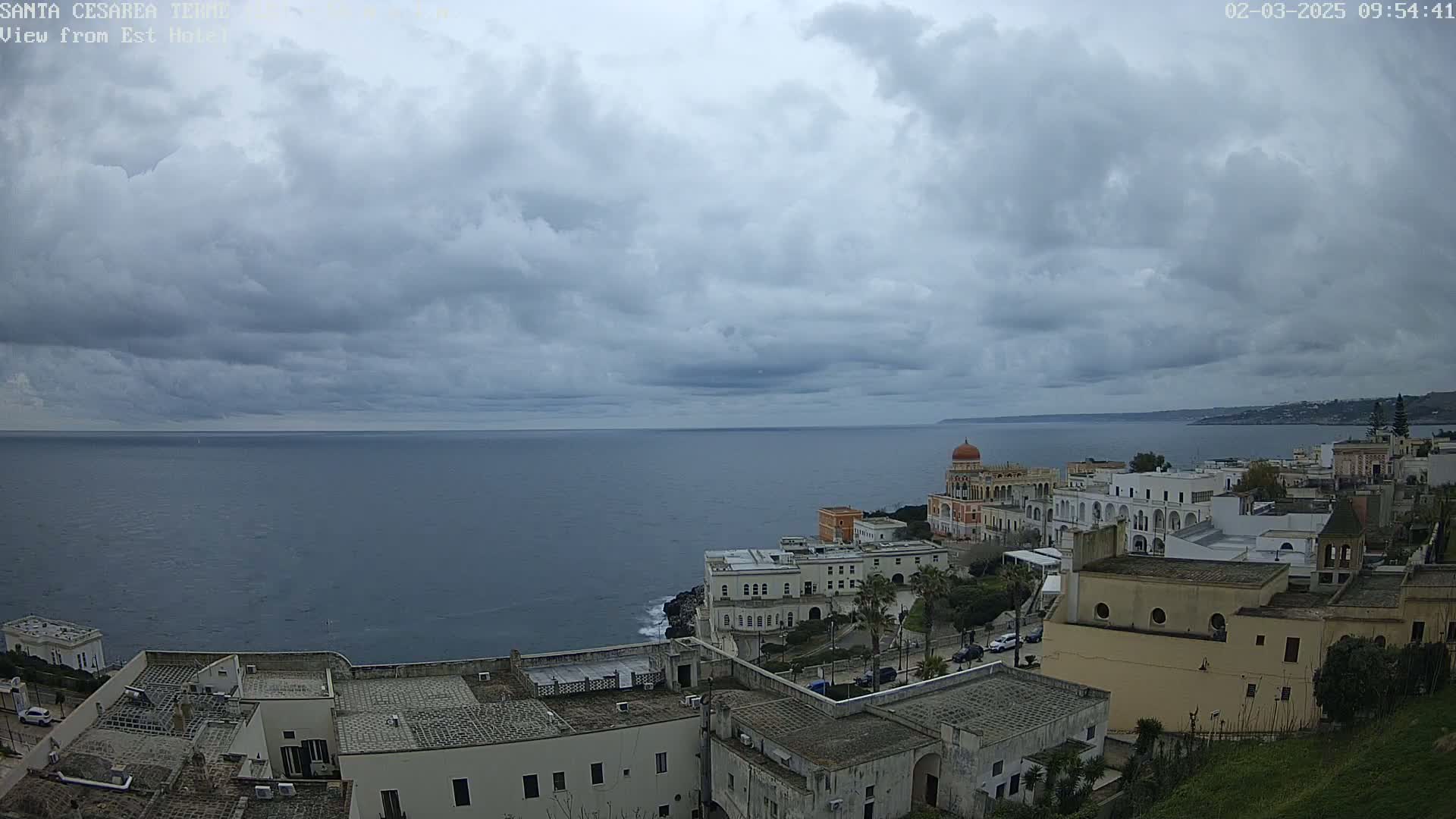 A coastal town with white buildings and a domed structure overlooks a calm sea under a cloudy sky.