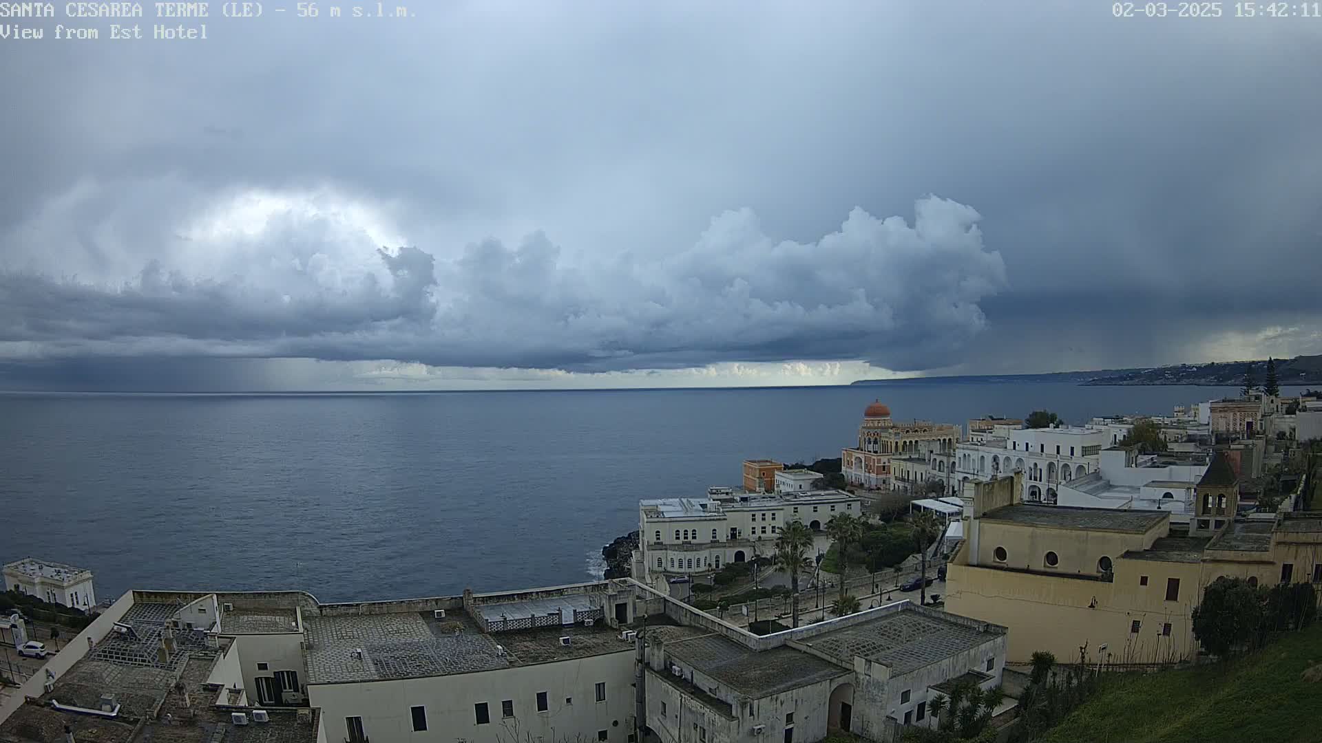 A coastal town with white buildings is situated below a dark, stormy sky over a calm sea.