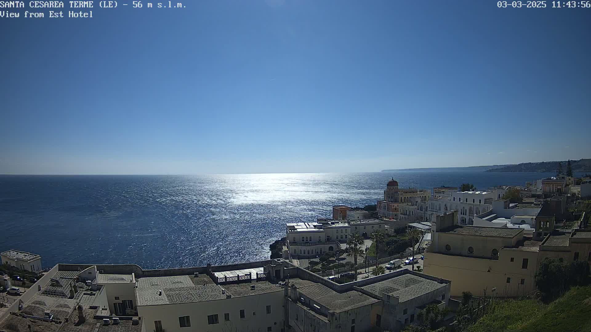 A coastal town with white buildings overlooks a calm, sunlit sea under a clear blue sky.