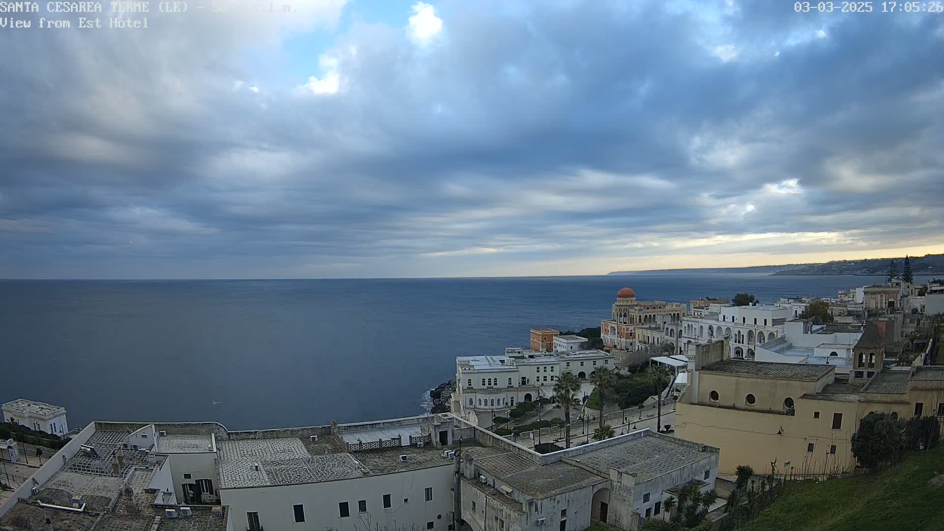A coastal town with white buildings and a domed structure overlooks a calm, dark-blue sea under a mostly cloudy sky.