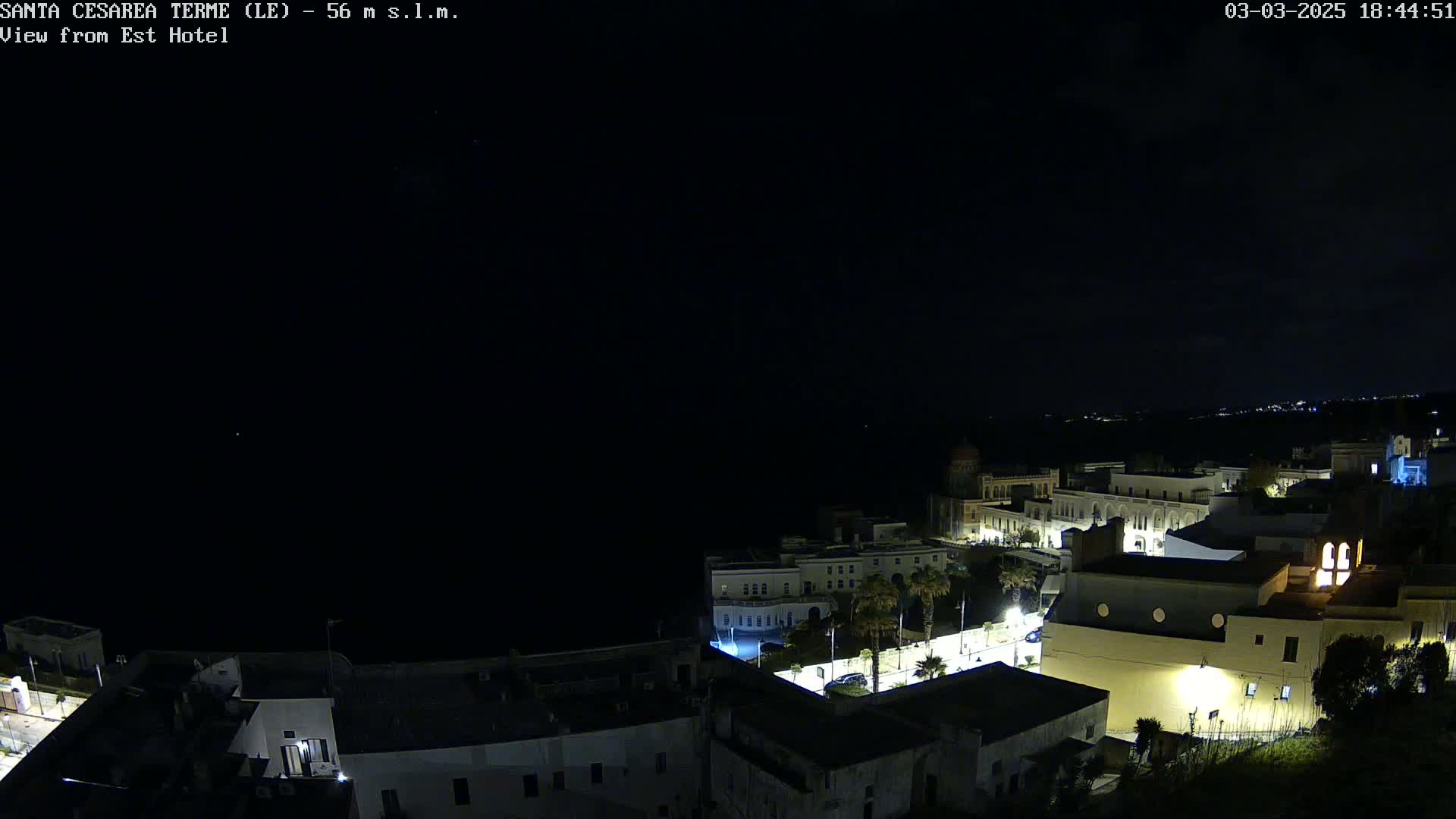 A nighttime, aerial view of a coastal town with dimly lit buildings and a dark sea under a clear sky.