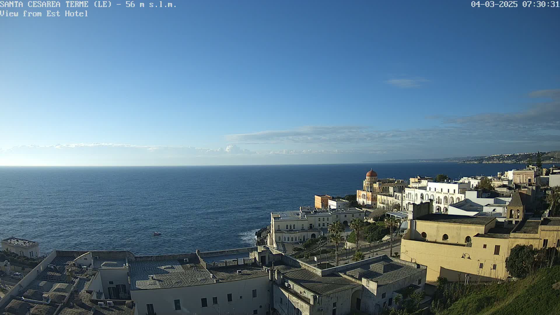 A coastal town with white buildings and a domed structure overlooks a calm blue sea under a clear blue sky.