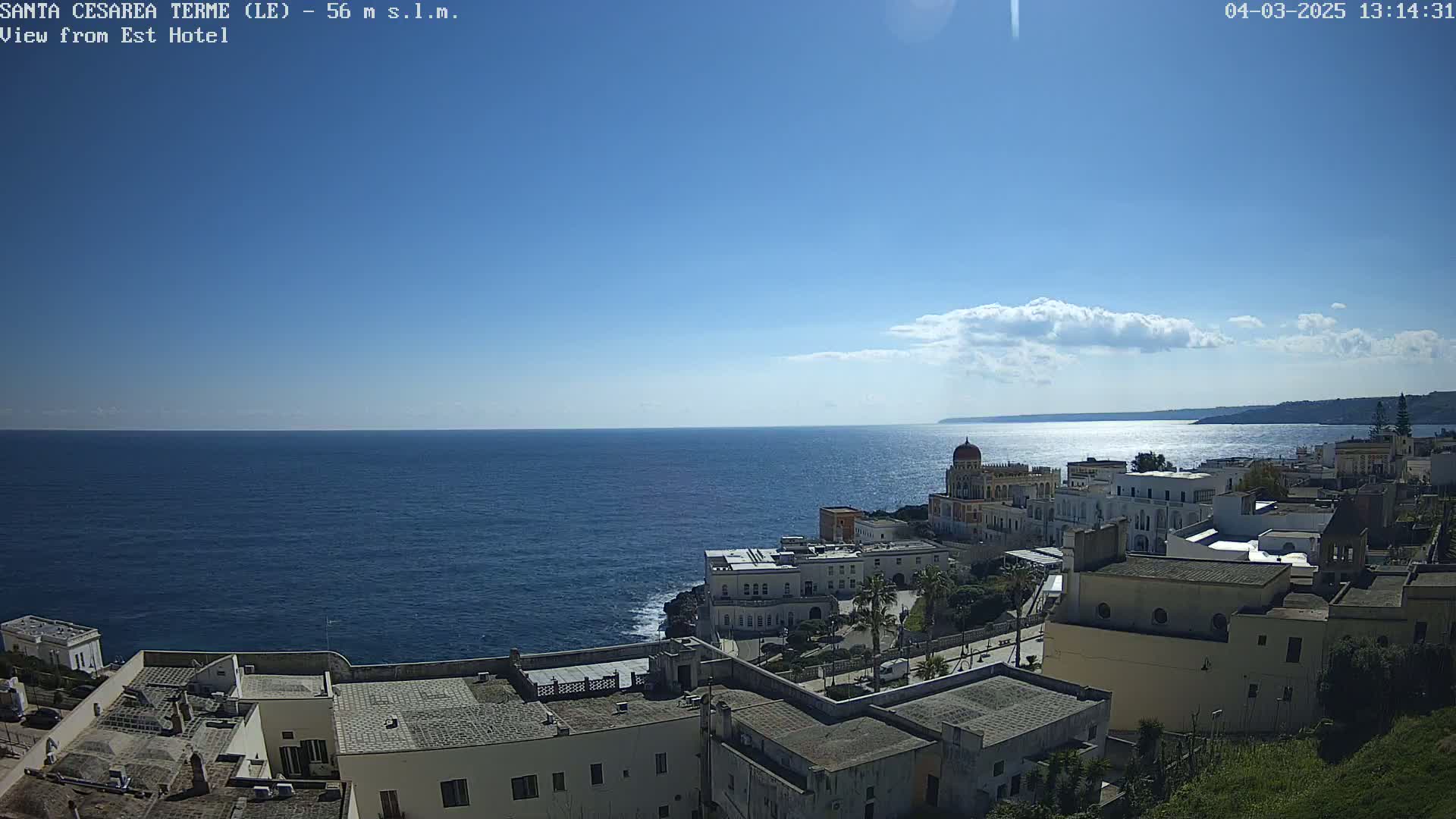 A coastal town with white buildings overlooks a calm blue sea under a mostly sunny sky with a few scattered clouds.