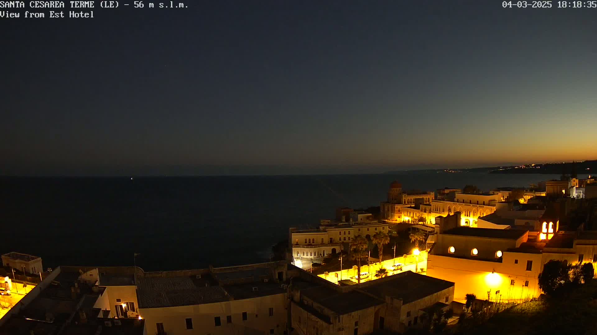 A coastal town at night is illuminated, with a dark ocean in the background under a twilight sky.