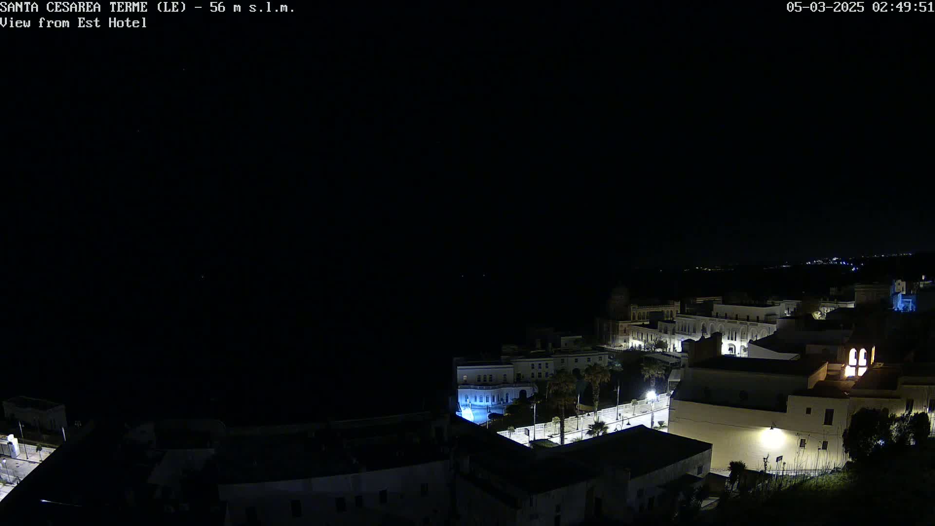A nighttime, high-angle view shows a coastal town with illuminated buildings and streets under a clear, dark sky.