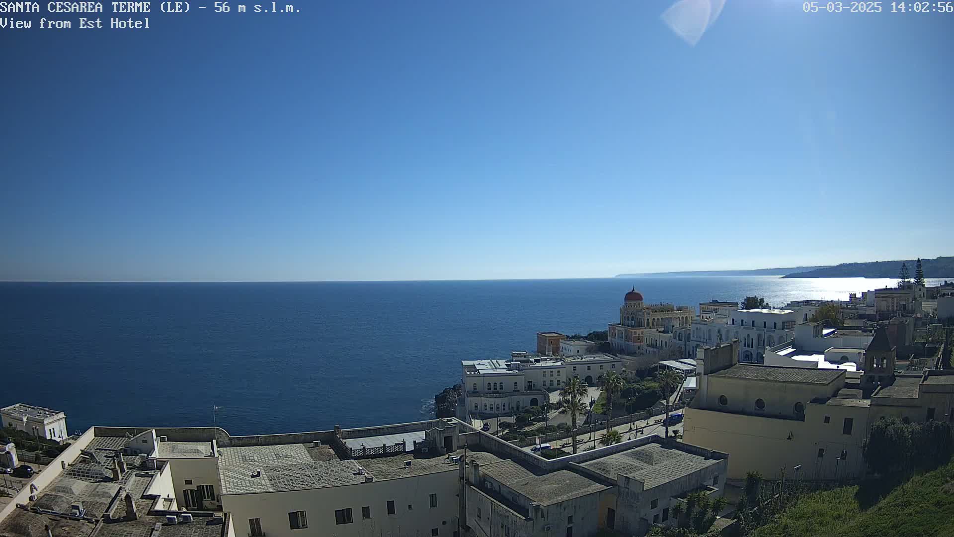 A coastal town with white buildings overlooks a calm, blue sea under a clear, sunny sky.