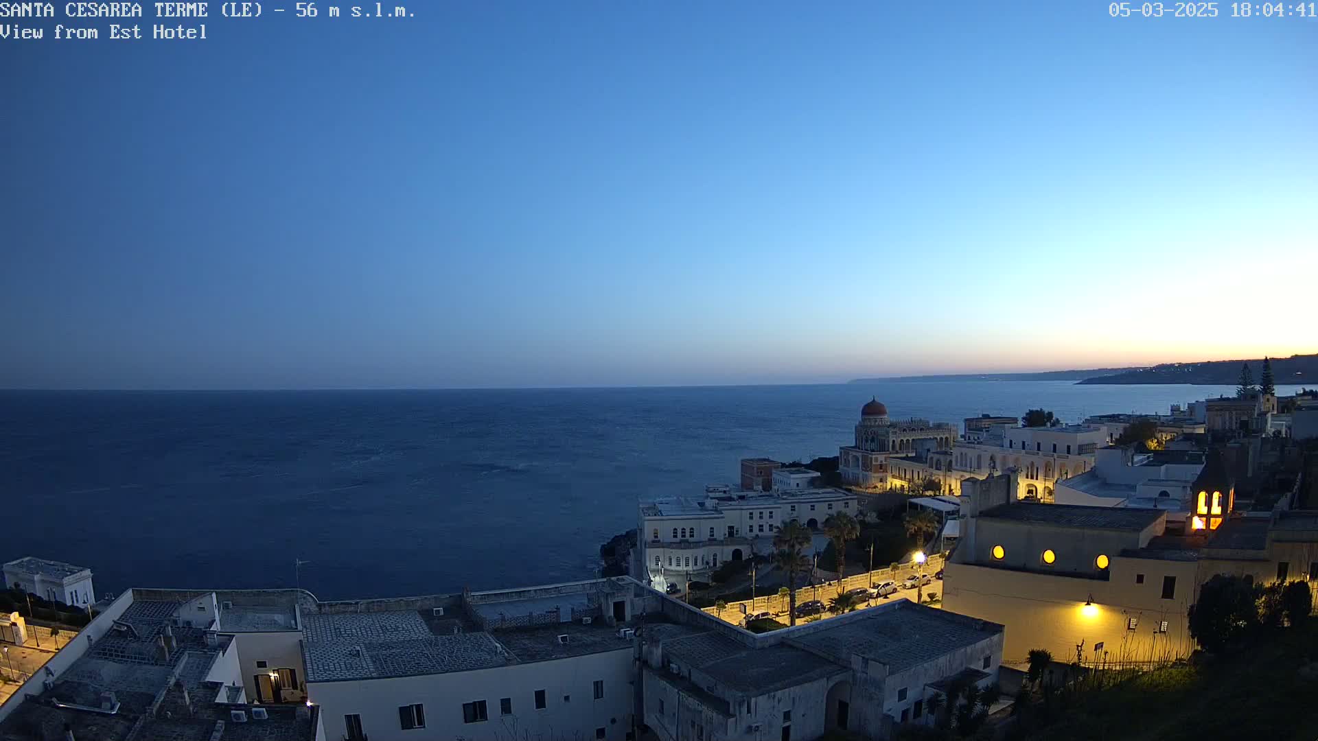 A coastal town at twilight, with calm dark blue water extending to the horizon under a clear, blue-tinged sky.
