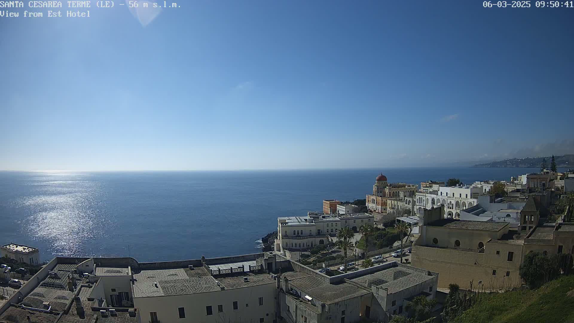 A coastal town with white buildings and a domed structure overlooks a calm, blue sea under a clear, sunny sky.