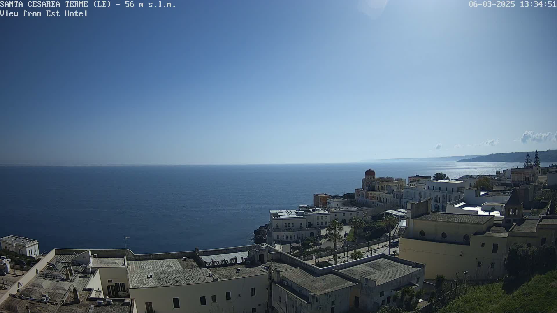A coastal town with white buildings overlooks a calm, blue sea under a clear, sunny sky.