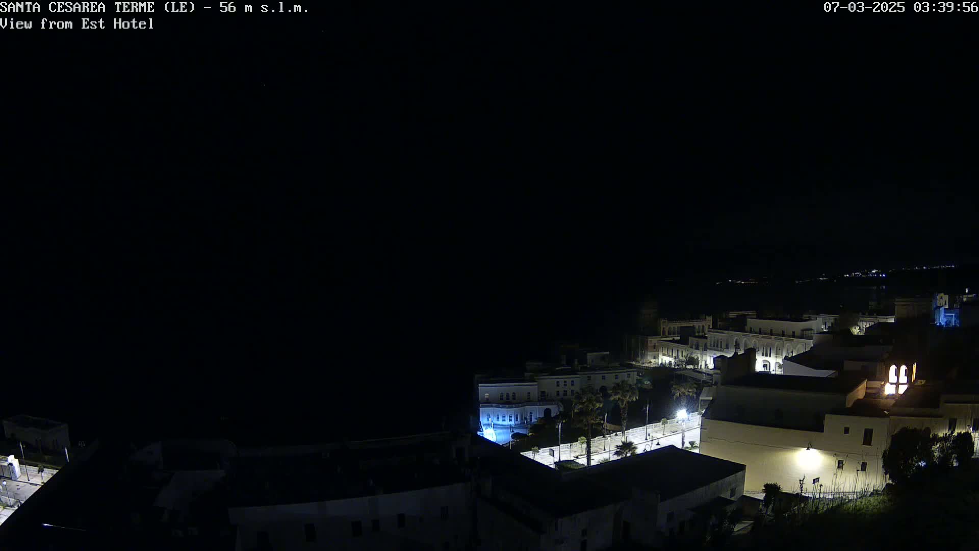 A nighttime, aerial view of a coastal town with buildings illuminated, under a clear, dark sky.