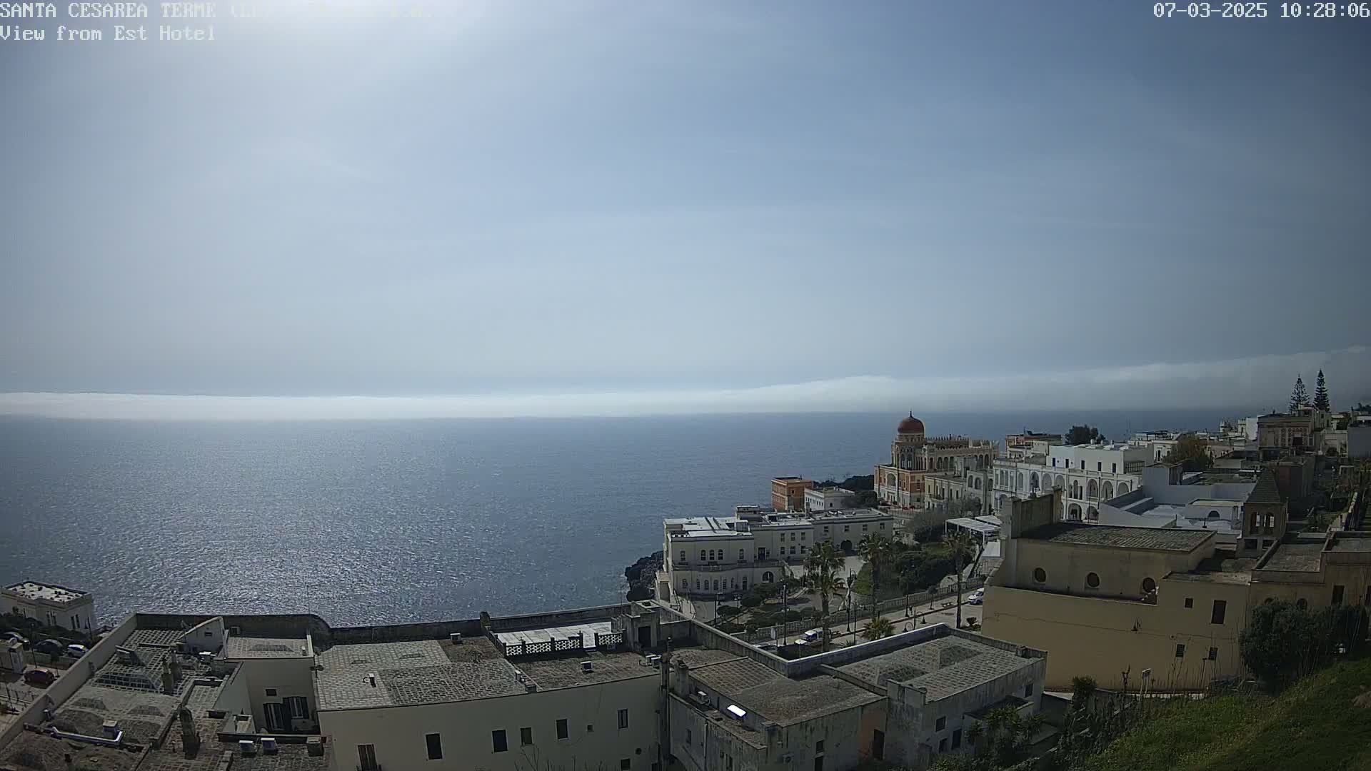 A coastal town with white buildings overlooks a calm sea under a hazy sky.