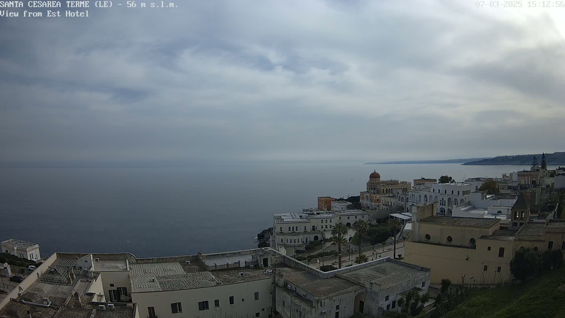 A coastal town with white buildings and a prominent domed structure overlooks a calm, gray sea under a cloudy sky.