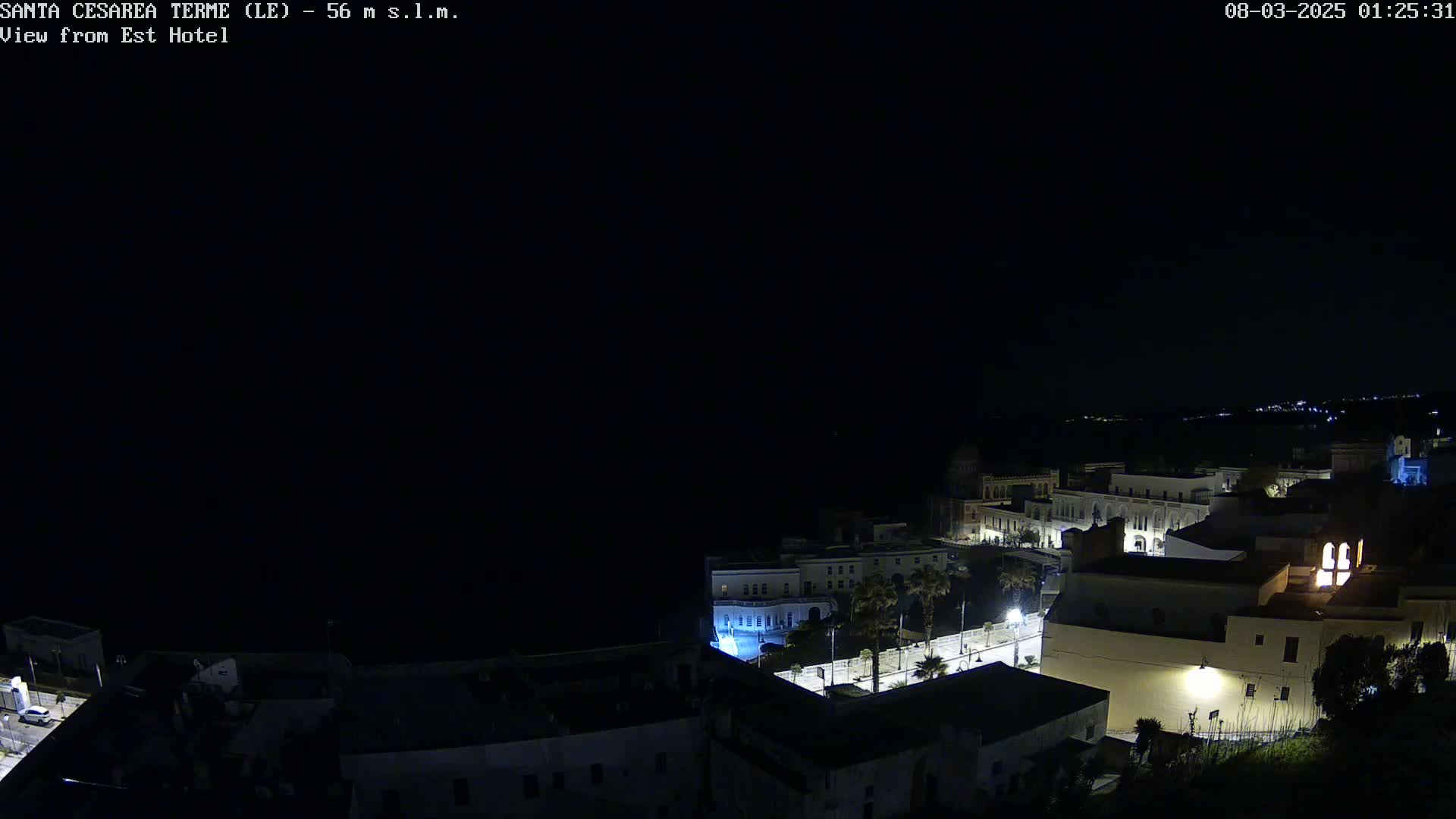 A nighttime view of a coastal town shows buildings illuminated along a dark shoreline under a clear, dark sky.