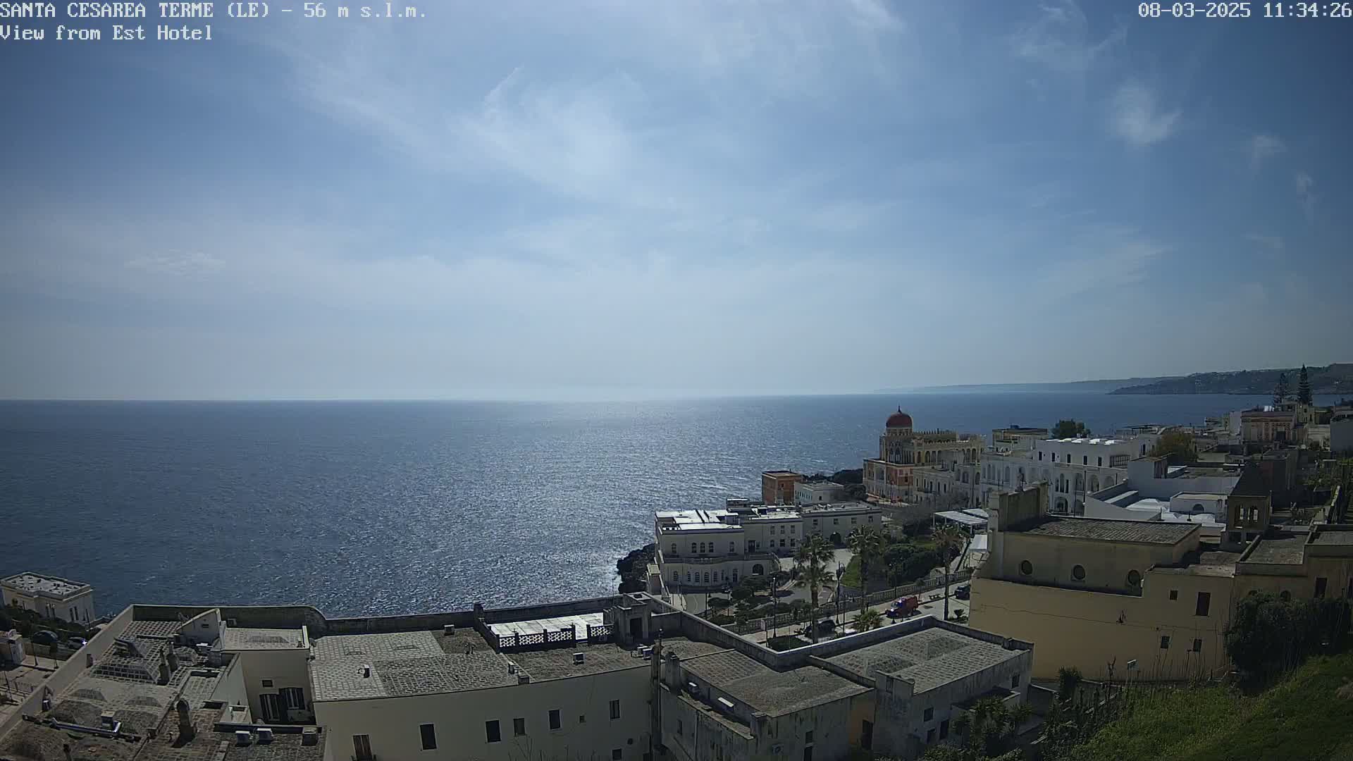 A coastal town with white buildings sits on a cliff overlooking a calm blue sea under a mostly sunny sky with a few scattered clouds.