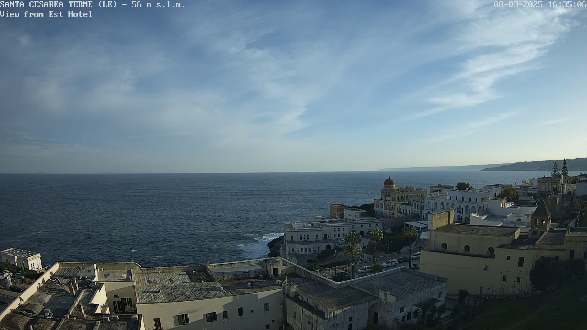 A coastal town with white buildings sits on a cliff overlooking a calm, dark blue sea under a mostly clear, light blue sky with a few wispy clouds.
