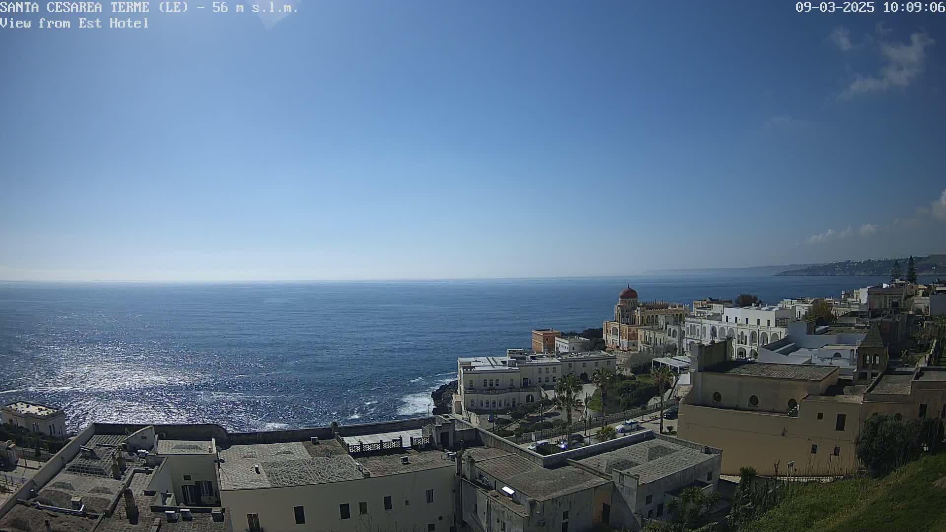 A coastal town with white buildings and a domed structure overlooks a calm blue sea under a clear blue sky.