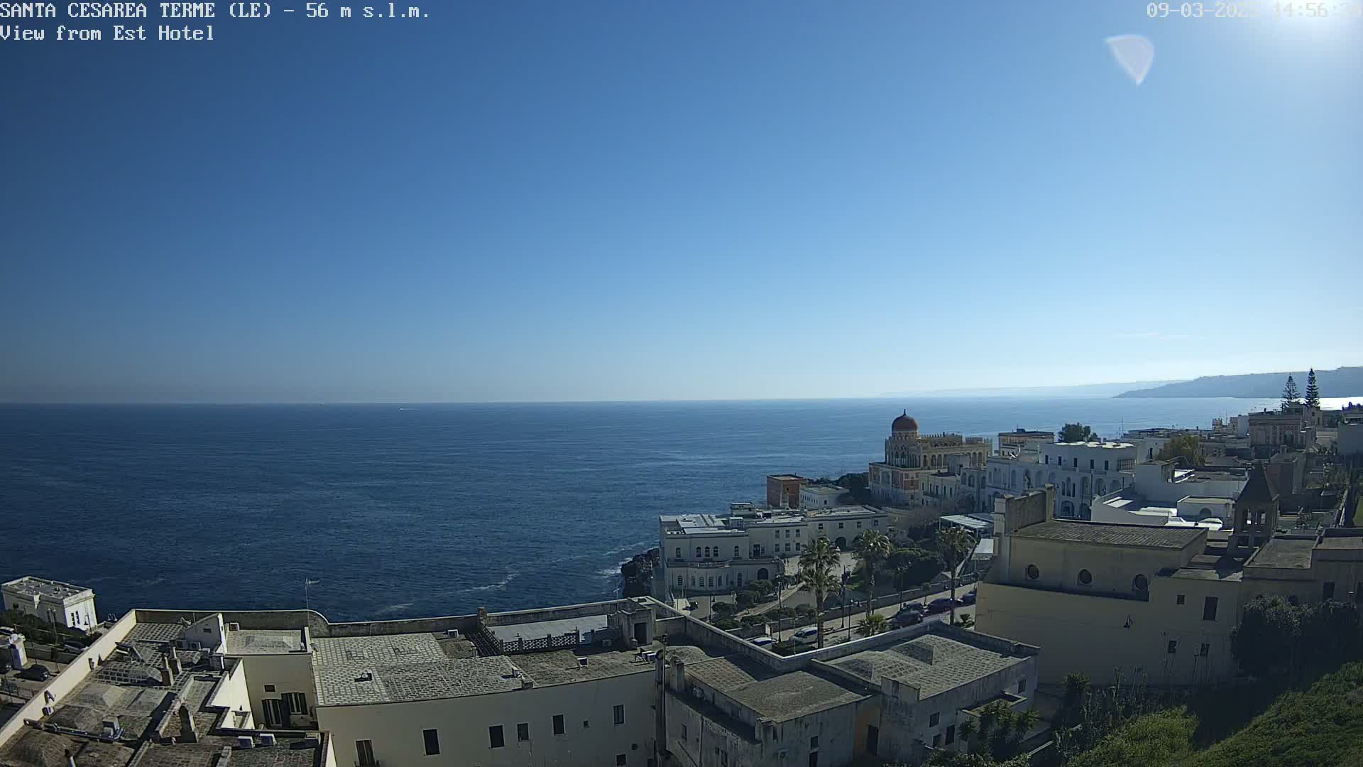 A coastal town with white buildings overlooks a calm, dark-blue sea under a clear, bright blue sky.