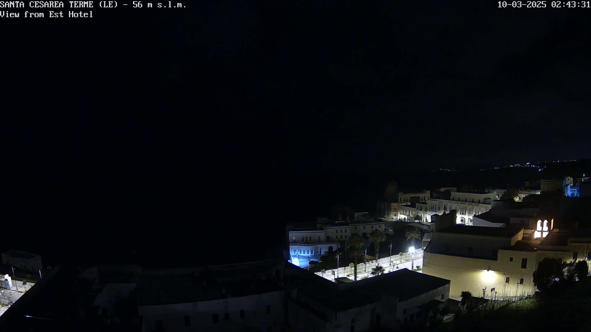 A nighttime, aerial view of a coastal town with buildings illuminated, overlooking a dark sea under a clear sky.