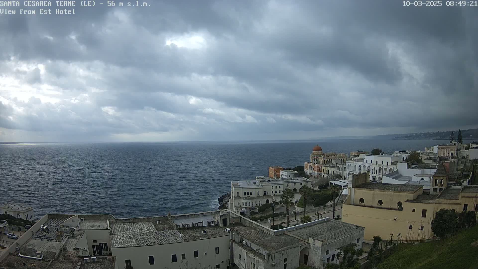 A coastal town with white buildings sits on a cliff overlooking a dark blue sea under a cloudy sky.