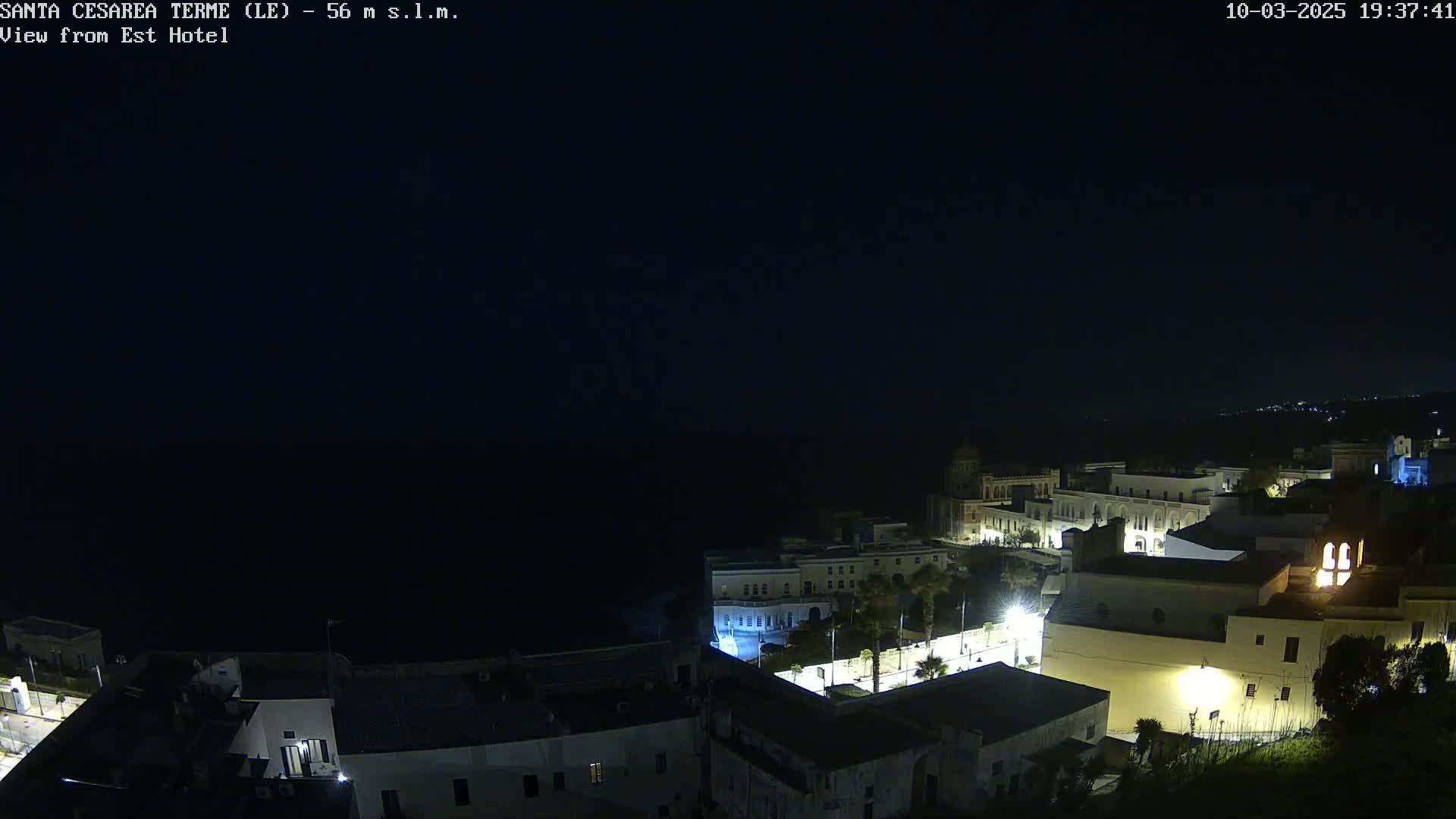 A nighttime coastal town is illuminated, with a dark ocean visible in the background under a clear, dark sky.