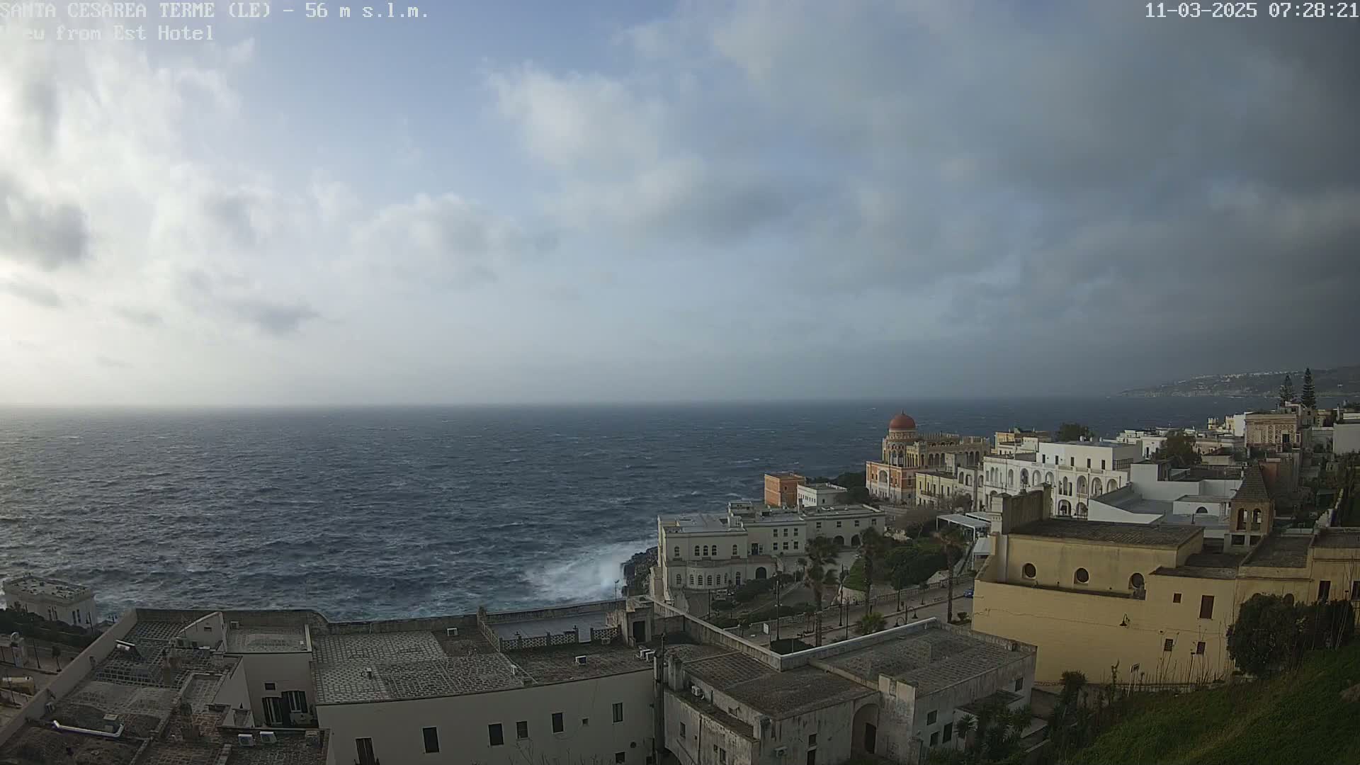 A windy, cloudy day overlooks a coastal town with white buildings and a domed structure,  with a rough sea crashing against the rocky shore.