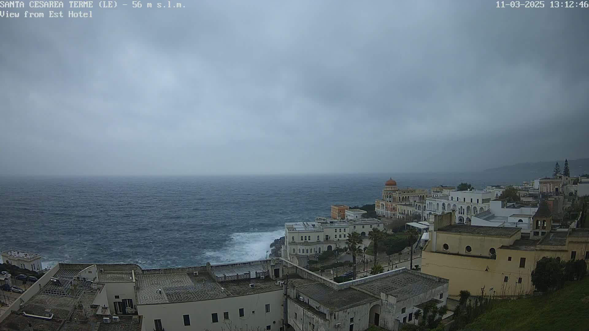 A coastal town with white buildings sits on a cliff overlooking a choppy sea under a gray, overcast sky.