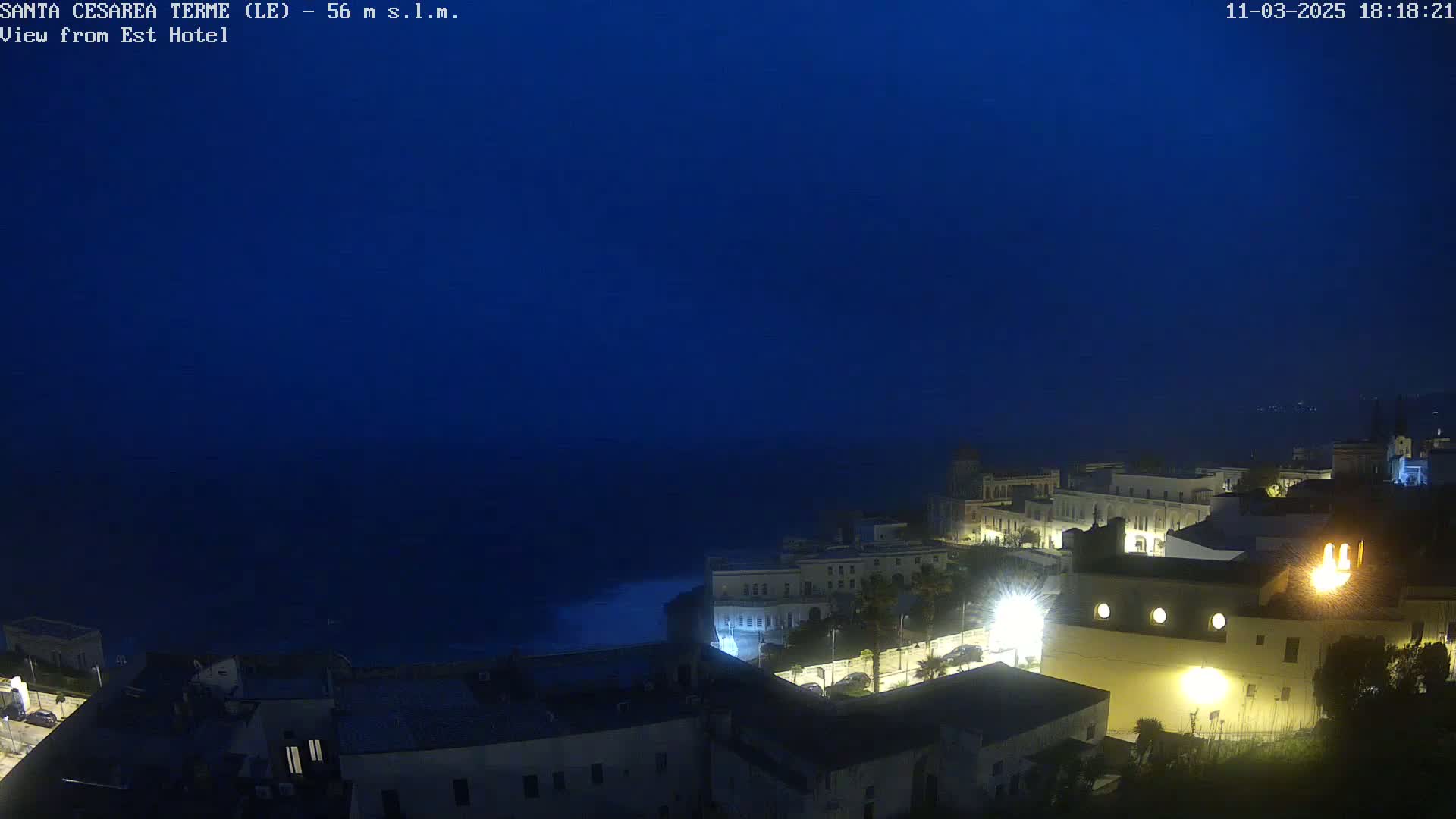A nighttime coastal town is dimly lit under a dark blue sky, with the dark sea visible in the background.