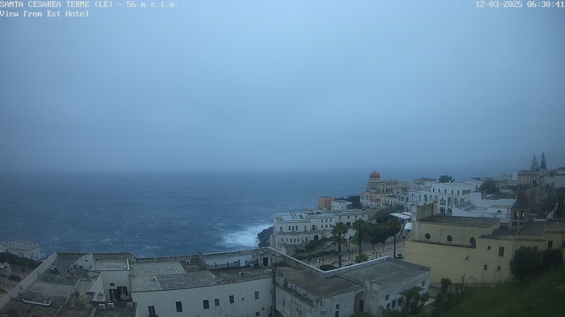 A coastal town with white buildings sits on a cliff overlooking a choppy, gray sea under an overcast sky.