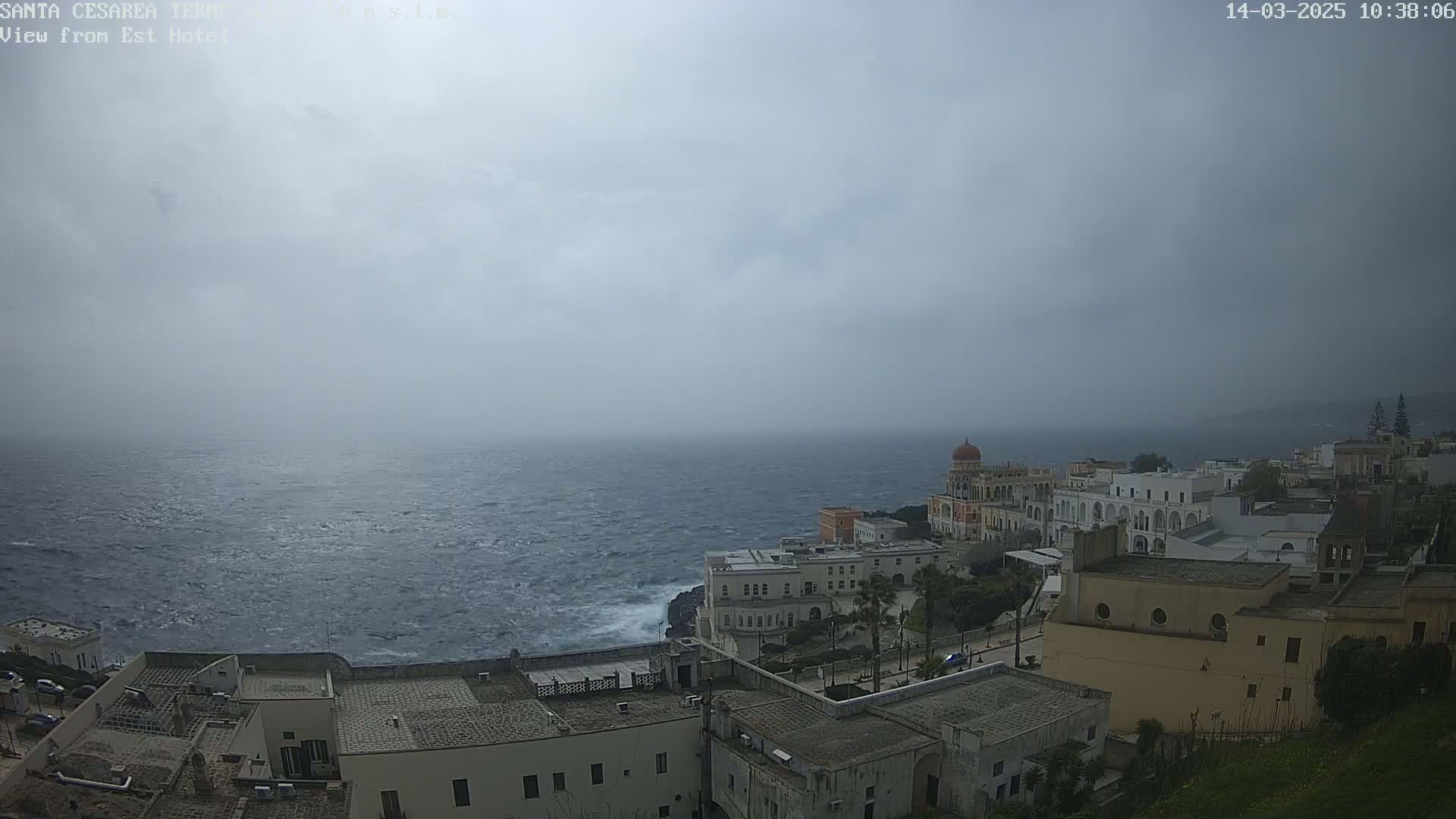 A coastal town with white buildings sits on a cliff overlooking a choppy sea under an overcast sky.
