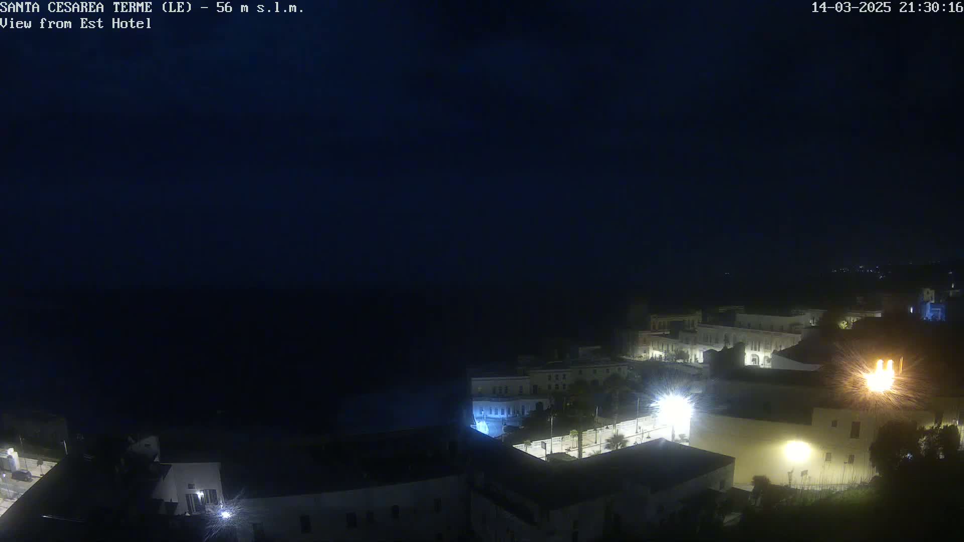 A nighttime, dark view of a coastal town with dimly lit buildings and a dark sea in the background.
