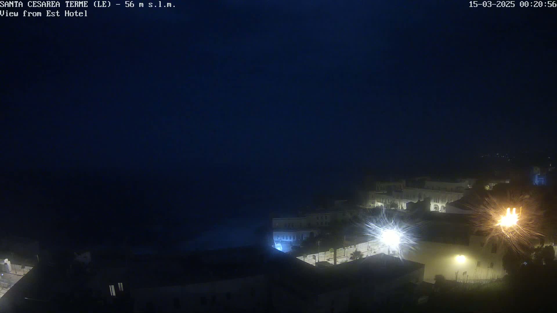 A nighttime, dark-blue coastal town is dimly lit with a few bright lights, overlooking a dark body of water under a clear night sky.
