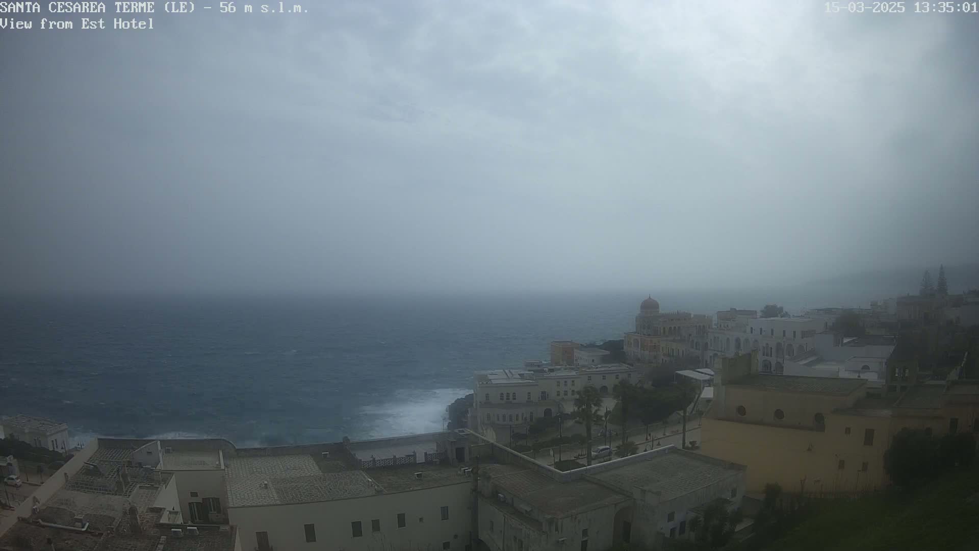 A hazy, overcast day reveals a coastal town with white buildings perched on a cliff overlooking a choppy sea.